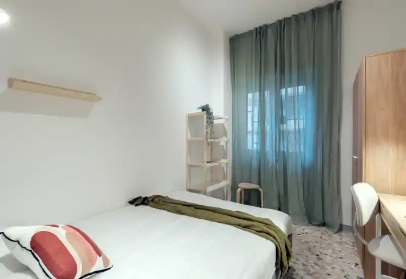 Dormitorio luminoso y minimalista con cama doble bien hecha, textiles suaves y una ventana alta con cortinas translúcidas que difuminan la luz natural.