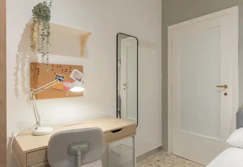 Rincón de dormitorio con escritorio de madera, lámpara de trabajo y silla junto al borde de la cama; tonos neutros, corcho para notas y espejo de cuerpo entero que aportan funcionalidad y estilo.