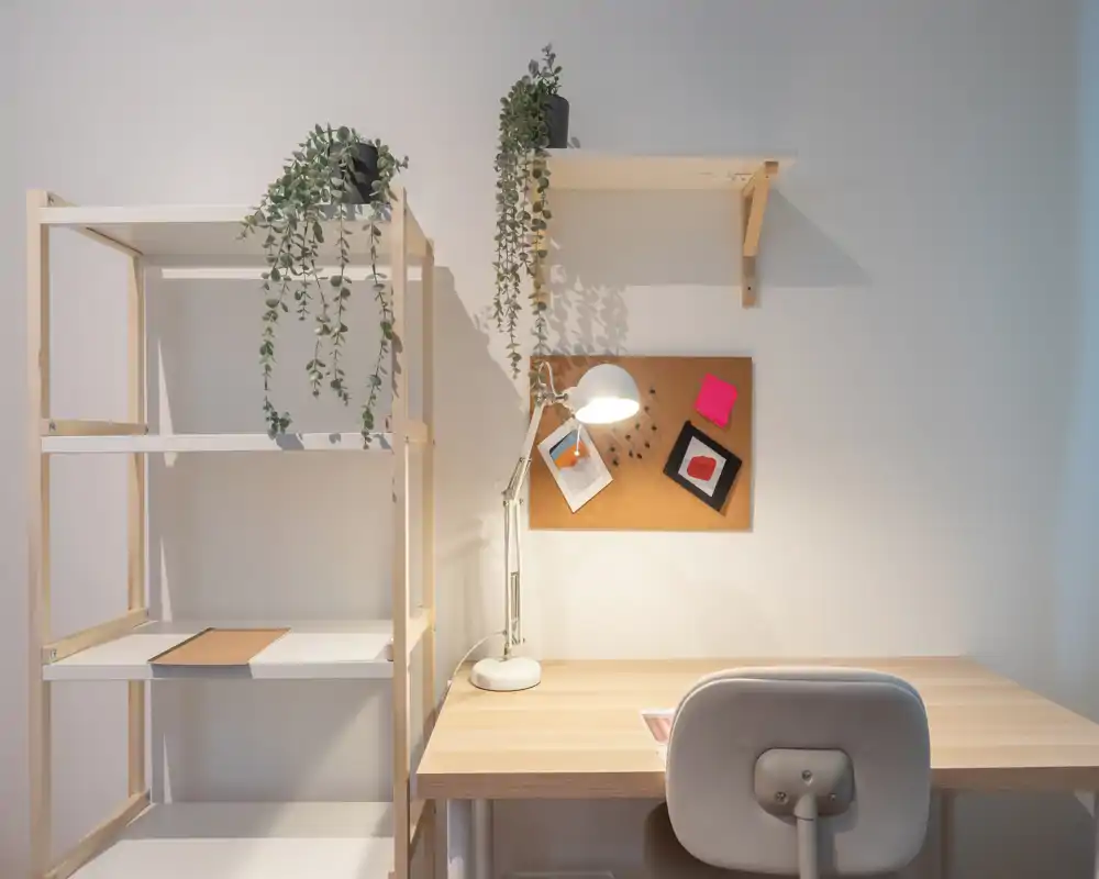 Angolo scrivania luminoso e minimale con tavolo in legno, lampada da lavoro e piante sospese; una bacheca in sughero sopra la scrivania crea un ordinato angolo home-office.