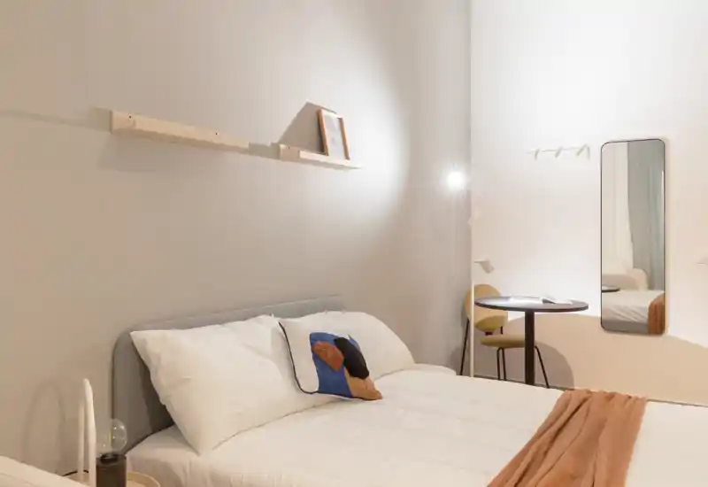 Camera da letto luminosa e minimal con un letto matrimoniale ben rifatto, cuscino decorativo e una coperta morbida. La stanza ha mensole semplici, una lampada da terra e uno specchio a parete che creano un'atmosfera calma e contemporanea.