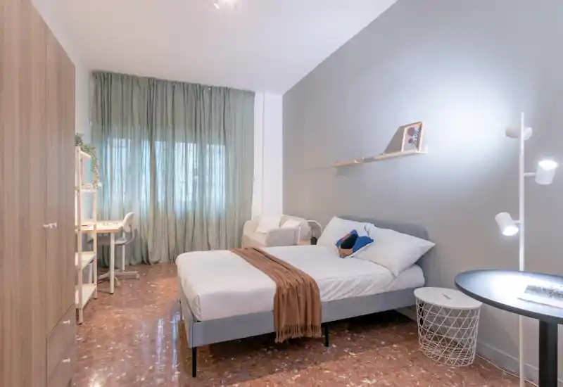 Camera da letto luminosa e ben arredata con letto completo, tessili morbidi e angolo con sedia/scrivania vicino alla finestra — perfetta per annunci che puntano su comfort e funzionalità.