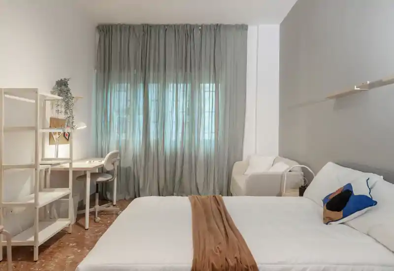 Camera da letto luminosa e curata con un ampio letto, biancheria morbida e una piccola scrivania vicino alla finestra. Toni neutri e arredi minimal rendono l’ambiente tranquillo e accogliente, ideale per affitti.