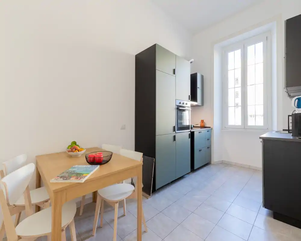 Cuisine moderne et lumineuse avec des éléments sombres, une table en bois et des chaises, et une grande fenêtre apportant une belle lumière naturelle—idéal pour les repas.