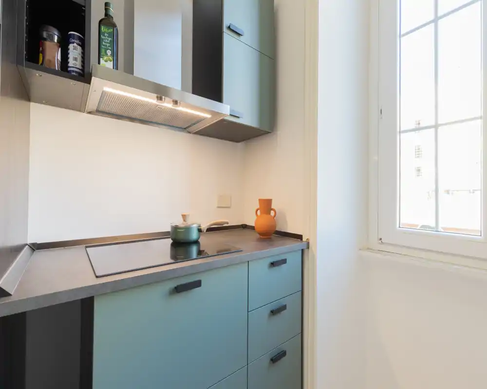 Cuisine compacte moderne avec plaque à induction intégrée, hotte et meubles bleu-vert clairs près d'une fenêtre lumineuse — propre, fonctionnelle et bien éclairée.