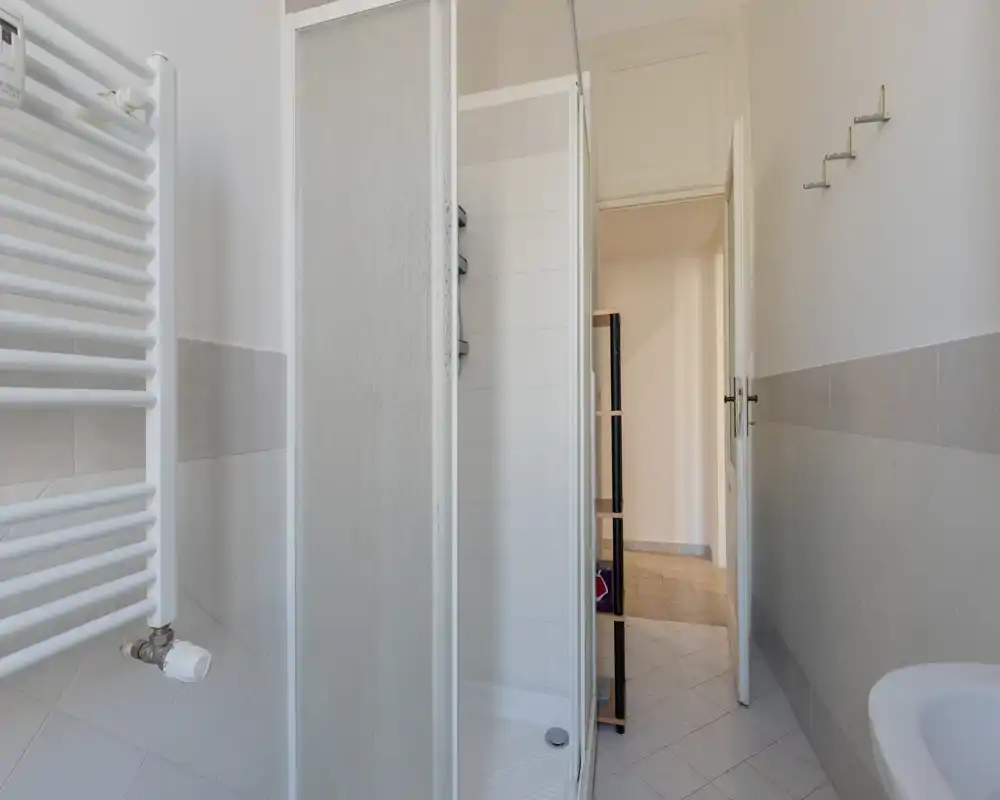 Petit salle de bain propre avec cabine de douche d'angle, radiateur porte-serviettes et lavabo visibles; carrelage neutre et lumière naturelle pour une atmosphère fraîche.