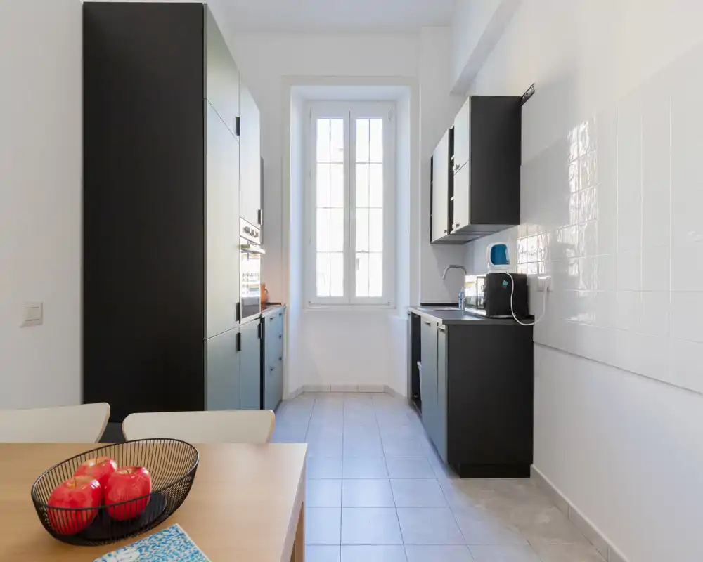 Cuisine en long moderne et lumineuse avec des placards foncés, carrelage et une fenêtre centrale ; une petite table avec des fruits apporte une touche chaleureuse.