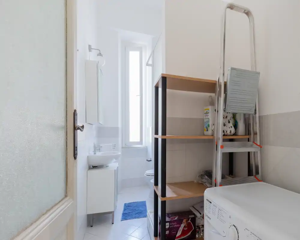 Salle de bain compacte et lumineuse avec lavabo, WC, fenêtre étroite et sol carrelé ; buanderie avec étagère et machine à laver au premier plan.