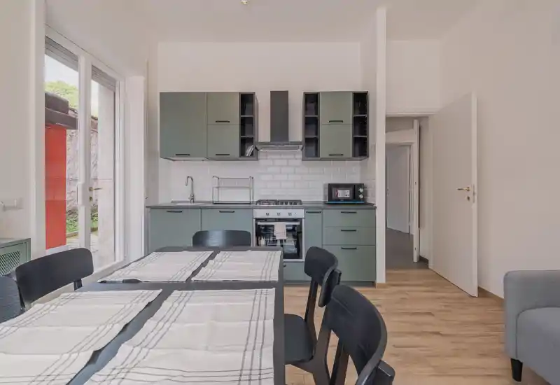 Cocina abierta moderna con armarios verde salvia, salpicadero de azulejos tipo metro y mesa de comedor en primer plano. La luz natural entra por una puerta acristalada, creando un área de cocina y comedor luminosa y acogedora.