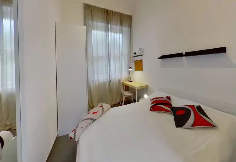 Dormitorio moderno y luminoso con una amplia cama blanca, cojines decorativos rojos y negros y un pequeño escritorio junto a la ventana. Paredes neutras, cortinas translúcidas y estantes minimalistas crean un espacio tranquilo y bien iluminado, ideal para descansar y trabajar.