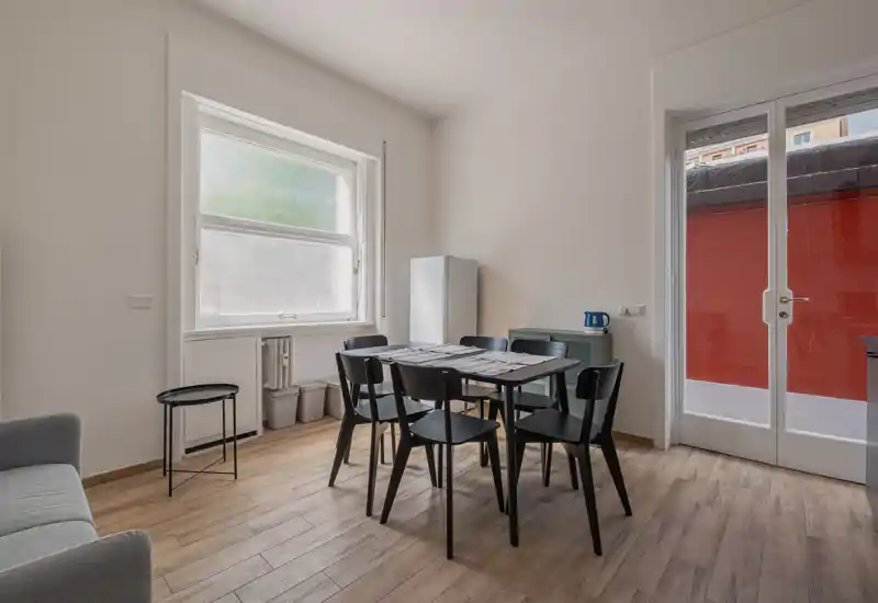 Zona de comedor moderna y minimalista con mesa rectangular y seis sillas negras junto a una gran ventana y puerta de cristal; luz natural abundante y suelo efecto madera que crean una sensación contemporánea y luminosa.