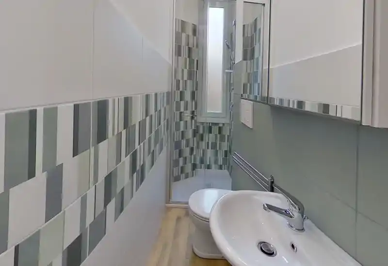 Compact, modern bathroom with a sink, wall-mounted toilet and a glass-enclosed shower. Decorative vertical tile banding and a frosted window provide natural light.