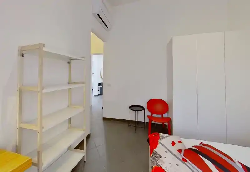 Rincón de dormitorio minimalista con cama parcialmente visible con edredón rojo y blanco, silla roja de acento, mesita redonda, armario blanco y estantería abierta. Luminoso, ordenado y apto para una sola persona.