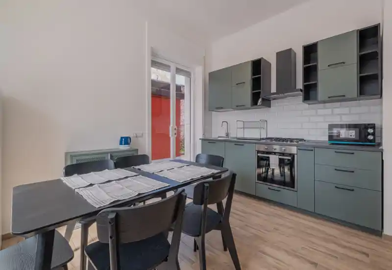 Cocina moderna abierta con muebles verdes mate, horno empotrado y placa de gas, y mesa negra para seis; la luz natural de la puerta de vidrio crea un ambiente acogedor.