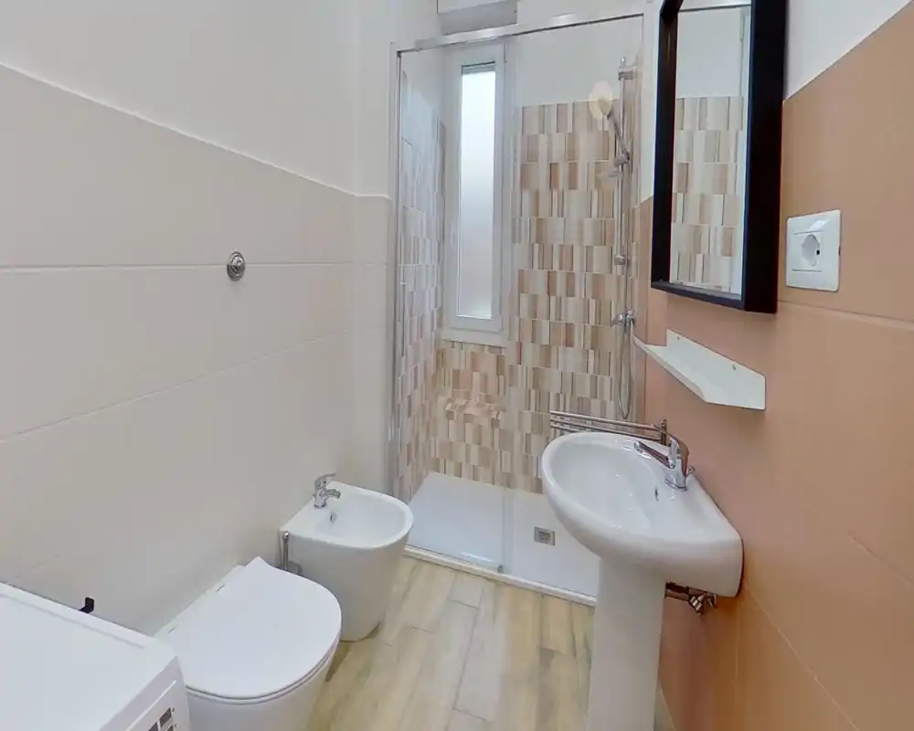 Baño moderno con cabina de ducha, lavabo de pie, bidé y WC; azulejos neutros y suelo efecto madera que crean un espacio limpio y luminoso.