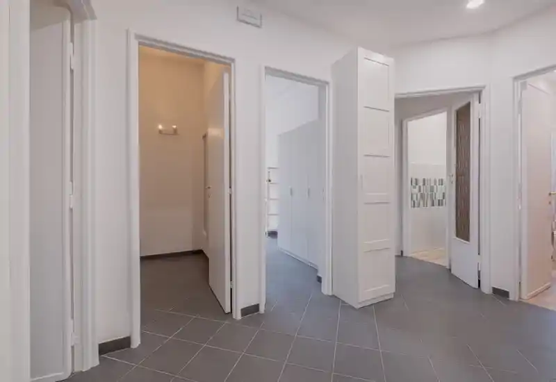 Entrée/couloir lumineux et minimaliste avec carrelage gris et plusieurs portes blanches menant aux pièces adjacentes ; rangement encastré visible.