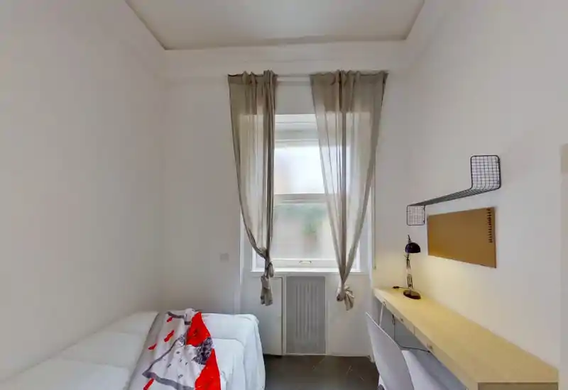 Chambre compacte et lumineuse avec un lit simple à gauche et un bureau long sous une lumière naturelle douce. Murs neutres, rideaux légers et déco minimaliste pour une pièce fonctionnelle et apaisante, parfaite pour une personne seule.