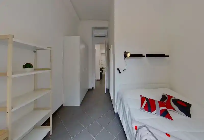 Chambre moderne et compacte avec lit blanc, coussins colorés, lampe de lecture murale, étagères ouvertes et armoire. Tons neutres et sol carrelé offrent un espace propre et confortable, idéal pour la location courte durée.