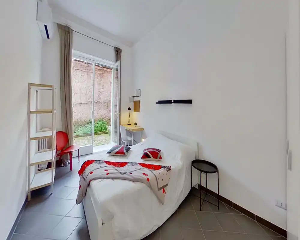 Bright, modern bedroom with a double bed, decorative pillows and a cozy throw. Large sliding glass door opens to a small outdoor area, and a compact desk by the window provides a workspace.