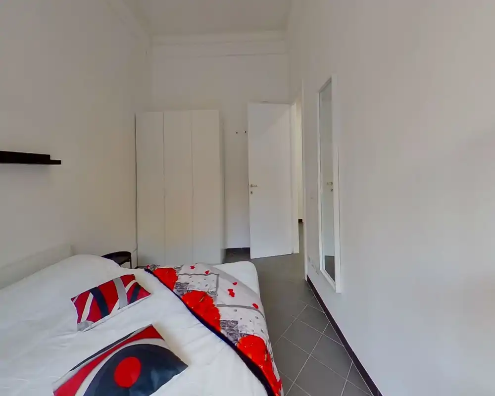 Minimal, well-lit bedroom with a double bed dressed in white linens and colourful cushions, a wardrobe and a full-length mirror. Neutral walls and tiled floor create a clean, contemporary look ideal for rental photos.
