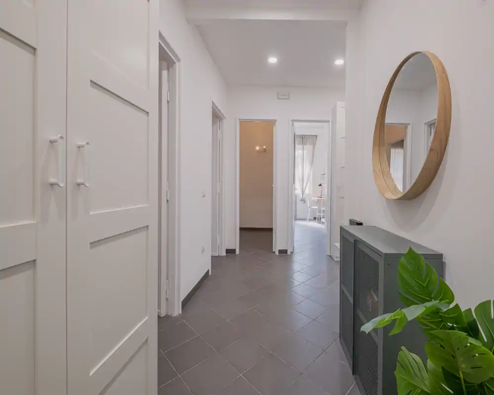 Bright, minimalist entrance corridor with tiled floor, built-in storage on the left, a console and round mirror on the right, and doors leading to other rooms.