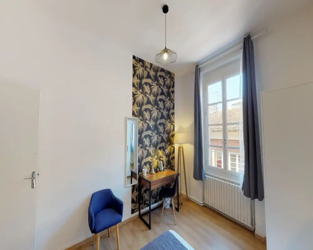 Angolo camera da letto luminoso e stiloso con parete decorativa, una piccola scrivania in legno e sedia accanto a una finestra che illumina naturalmente l’ambiente. La stanza ha una lampada da terra, uno specchio e una poltroncina, creando un'accogliente area lavoro nella zona notte.
