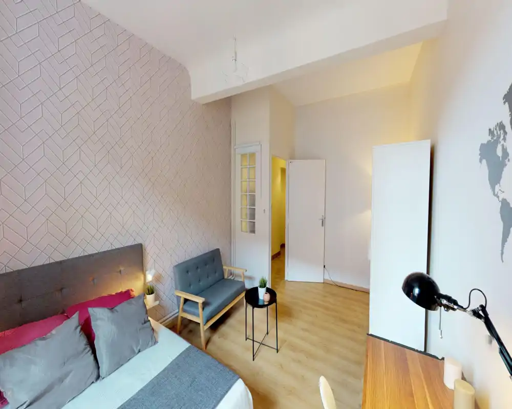 Spacious, well-lit bedroom with a comfortable double bed, patterned accent wall and a small seating area. Light wood floors and minimal modern furniture create a cozy, uncluttered feel ideal for rentals.