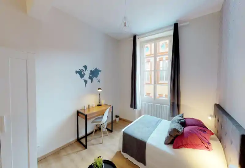Bright, modern bedroom with a comfortable double bed, crisp linens and decorative pillows. A wooden desk by the wall under a world-map decal and a window with long curtains provide a pleasant workspace and natural light.