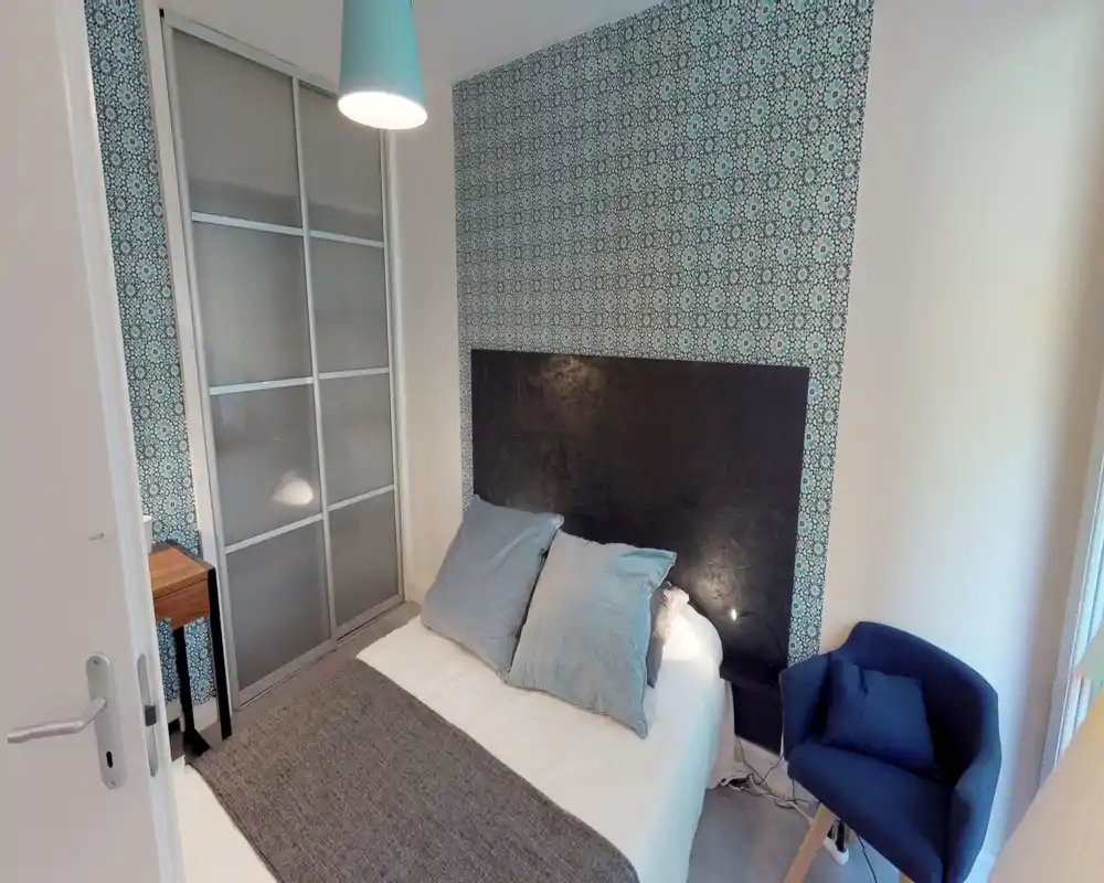 Camera da letto compatta con letto matrimoniale, parete decorativa a motivo, lampada da comodino e poltrona blu — luminosa e ben arredata, ideale per un annuncio.