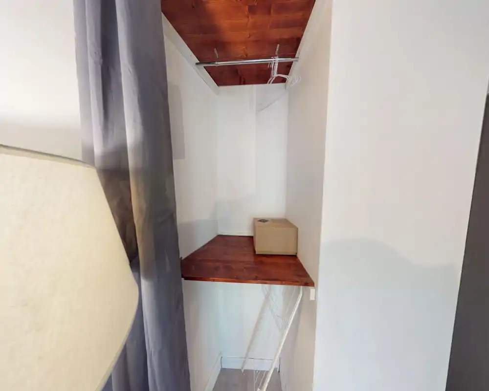Small built-in closet or storage nook with a wooden shelf, hanging rod and a few wire hangers; neutral white walls and a box on the shelf.