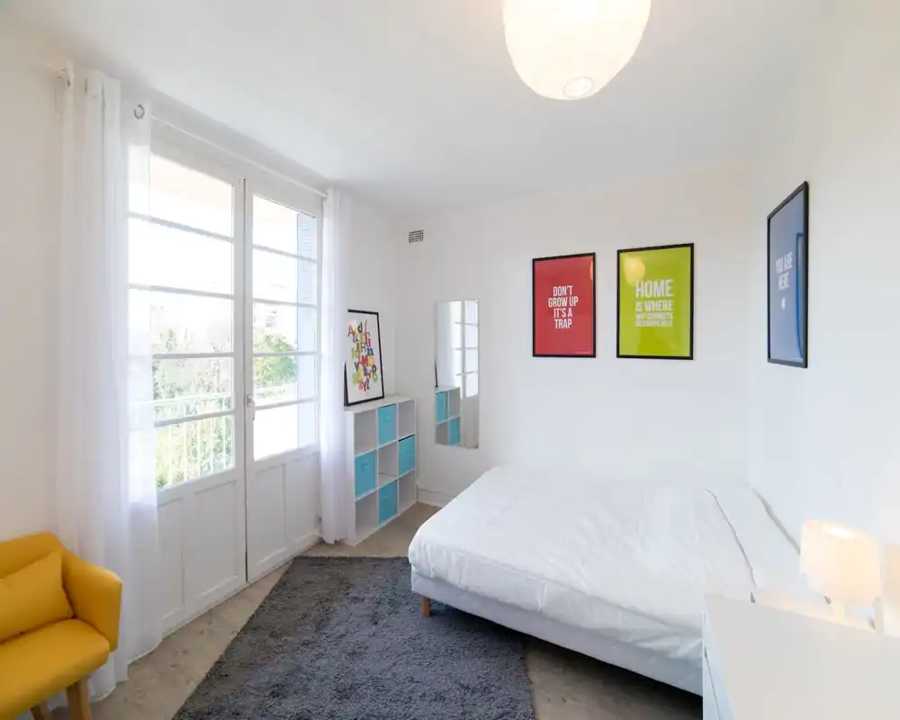 Bright, minimalist bedroom with a comfortable double bed, large window letting in abundant natural light, a soft rug and colorful framed prints that add personality.