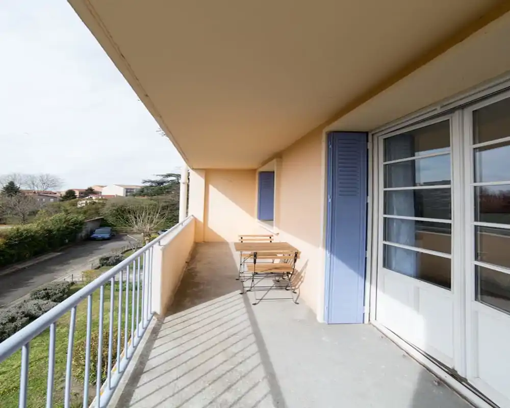 Sunny narrow balcony with metal railing, small foldable wooden table and chairs, and large glass doors opening to the interior — ideal for morning coffee with a neighborhood view.
