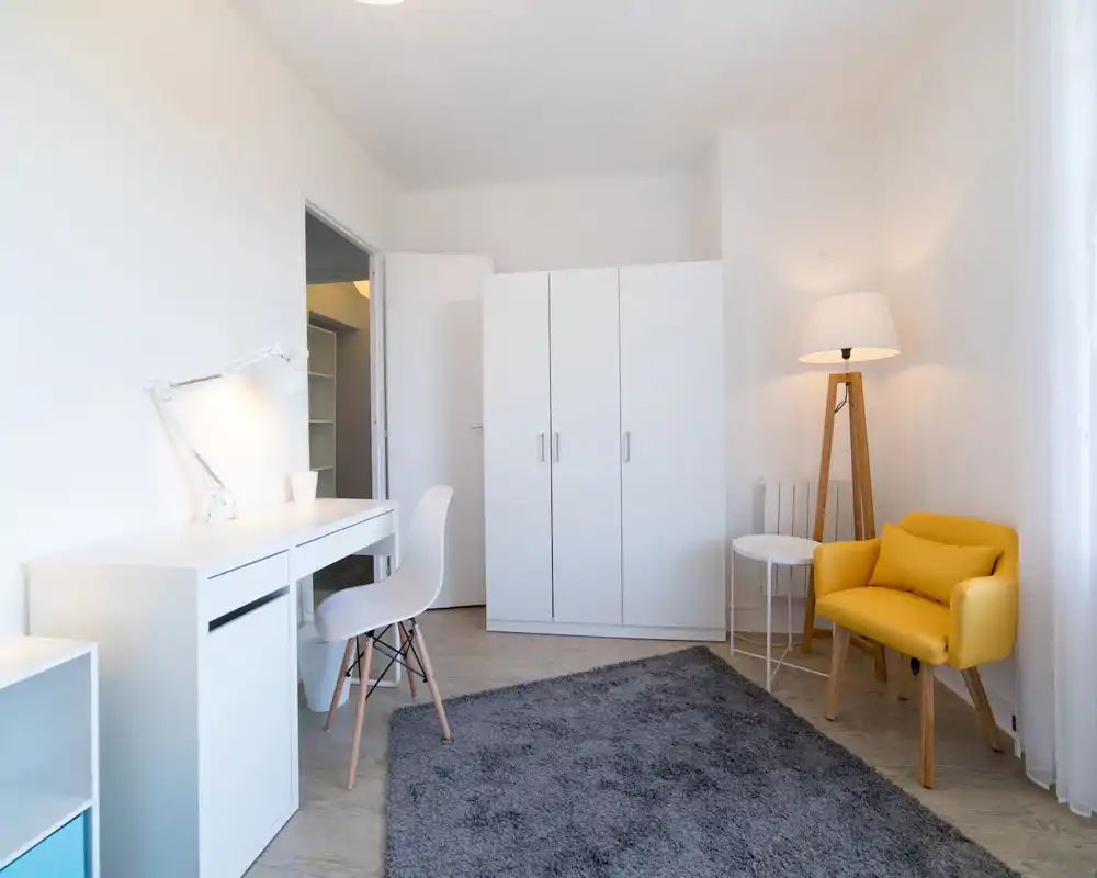 Bright, minimal bedroom with white furnishings, a wardrobe, desk and a yellow accent armchair. Large window light and neutral decor create a calm, airy space suitable for work and relaxation.