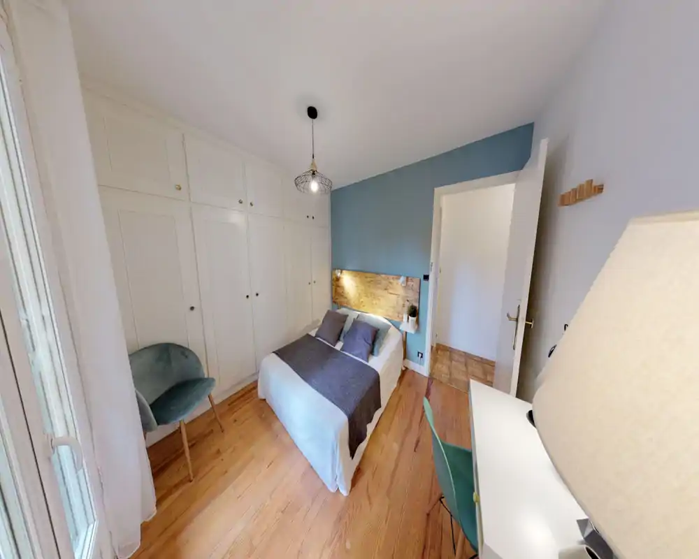 Camera da letto luminosa e di design con testiera in legno, letto matrimoniale con biancheria neutra e cuscini decorativi, armadi a muro e una poltroncina vicino alla finestra.