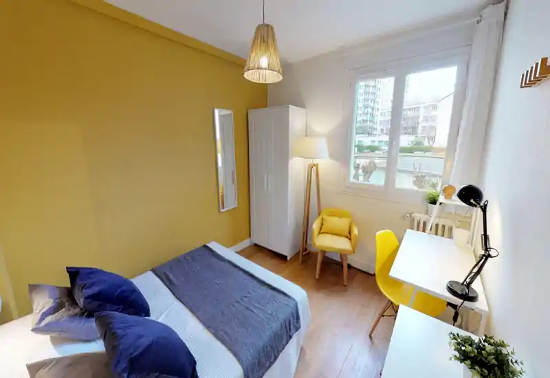 Bright, stylish bedroom with a large bed in the foreground, coordinated yellow accent wall and seating, a white wardrobe and desk by the window providing natural light — ideal for comfortable short or long stays.