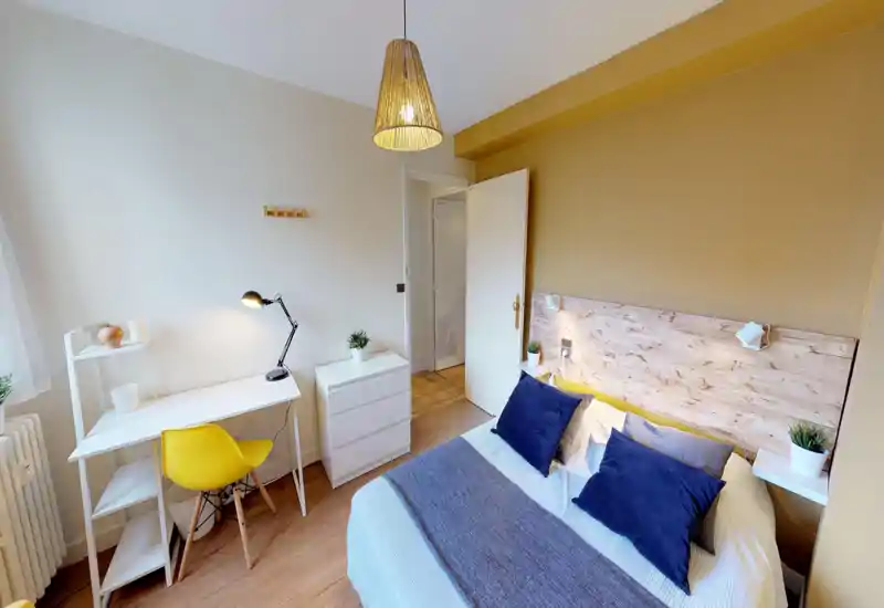 Bright, stylish bedroom with a comfortable double bed, navy cushions and a wooden headboard; includes a small desk with a yellow chair and natural light from the window.