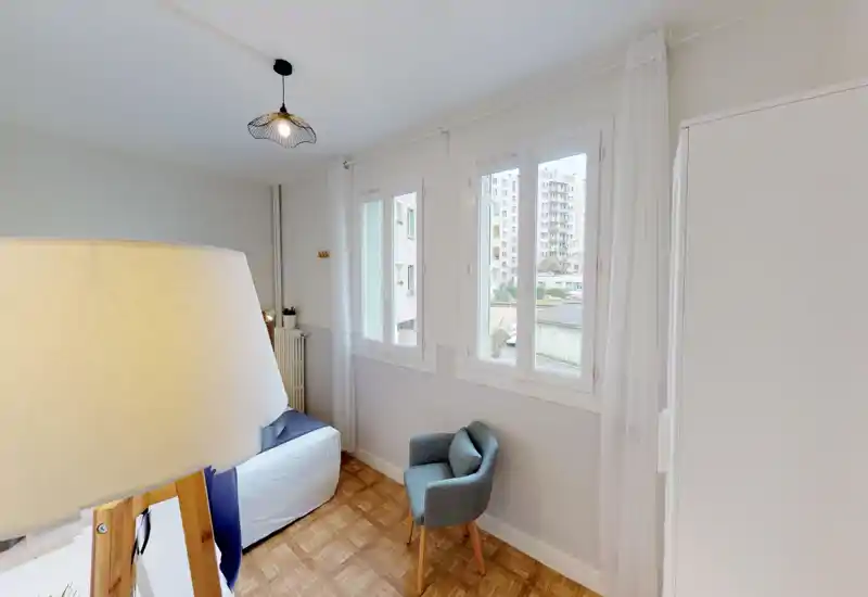 Bright, simply furnished bedroom with a partially visible bed, a cozy armchair and large windows providing natural light.