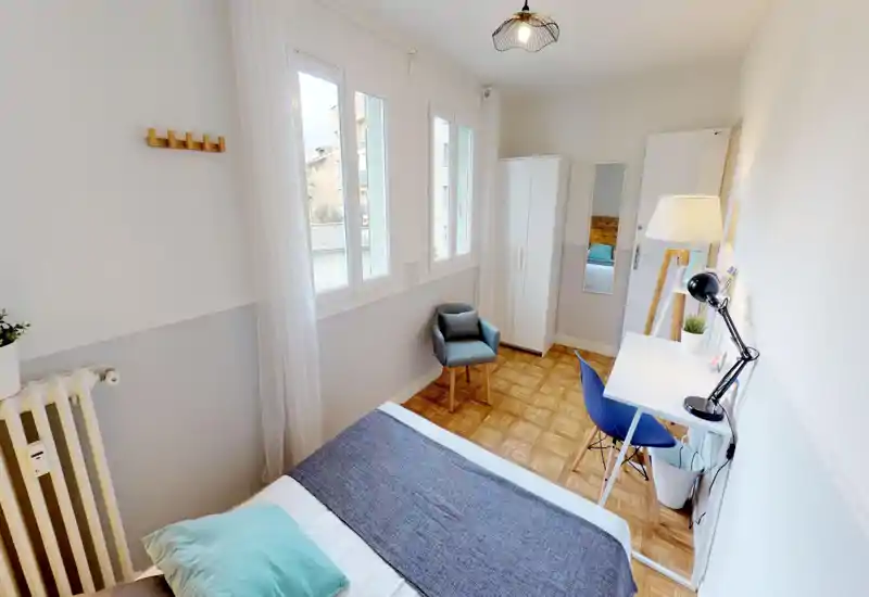 Bright, well-staged bedroom with a large window, cozy textiles and a small workspace. The bed is clearly visible and the room features a wardrobe, armchair and wooden parquet flooring.