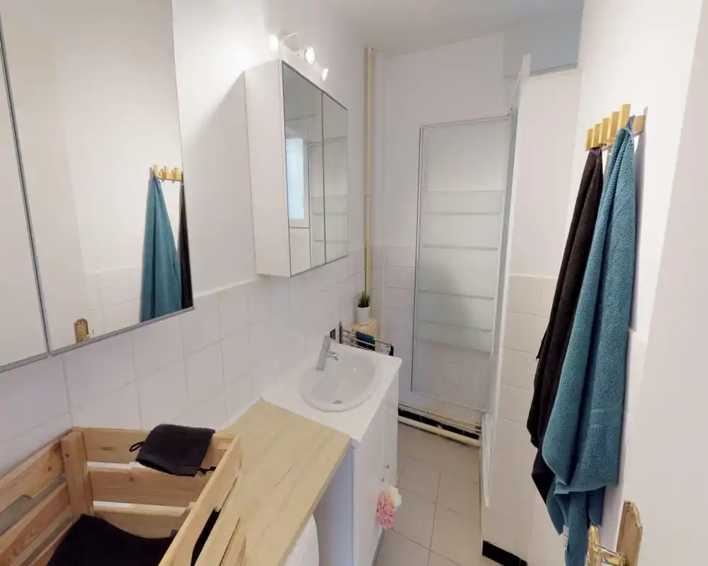 Bright, compact bathroom with a sink, mirrored medicine cabinet, tiled walls and a frosted shower screen; fresh towels and light wood accents give a clean, functional feel.