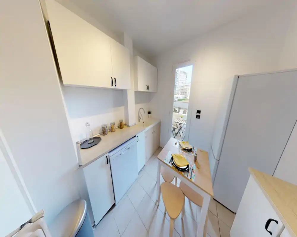 Compact modern kitchen with light wood countertops, white cabinets and a small dining table set for two. A glass door brings natural light and access to a small balcony.