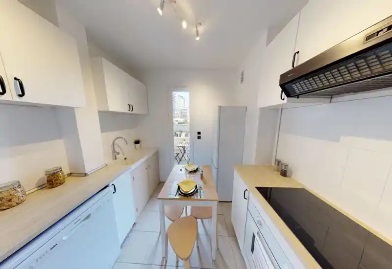 Cuisine lumineuse et compacte avec placards blancs, plans de travail effet bois, petite table centrale dressée pour deux et appareils modernes. Espace propre et accueillant avec lumière naturelle venant de la porte du balcon.