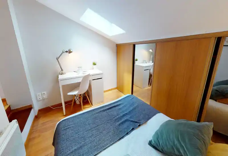 Chambre lumineuse et chaleureuse avec lit double au premier plan, petit bureau blanc et chaise sous une fenêtre de toit, et des placards coulissants en bois avec panneau miroir.