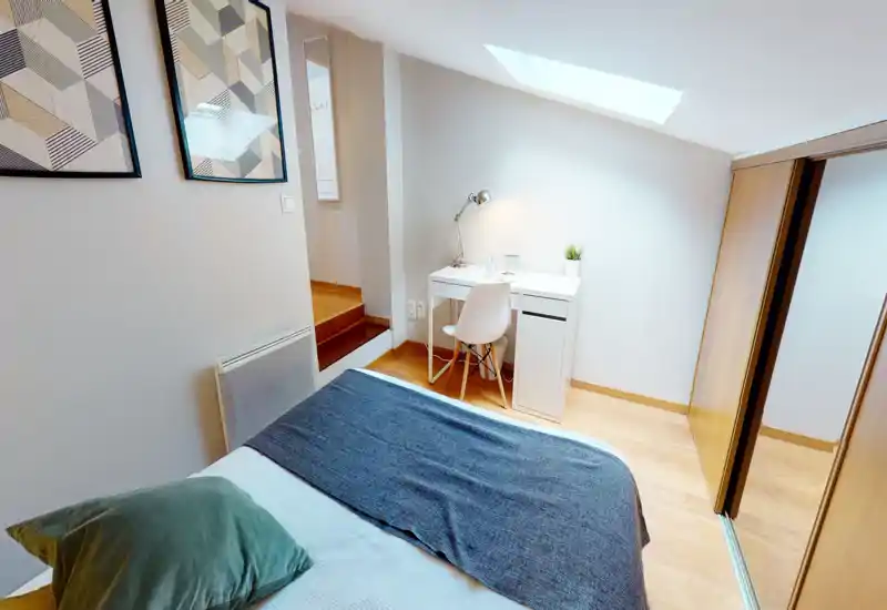 Chambre compacte et lumineuse avec un lit confortable au premier plan, un petit bureau blanc sous une fenêtre de toit et des placards coulissants. Décoration moderne et épurée avec un sol en bois et touches décoratives discrètes.