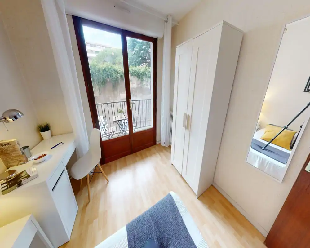 Bright, modern bedroom with large floor-to-ceiling window opening onto a small balcony, simple white wardrobe, desk and a neatly made bed with accent cushions.