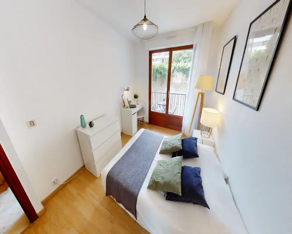 Bright, stylish bedroom with a double bed dressed in white linens, decorative cushions and a textured runner. Large glass doors open to a small balcony, and a compact desk and chest of drawers provide a functional work corner.