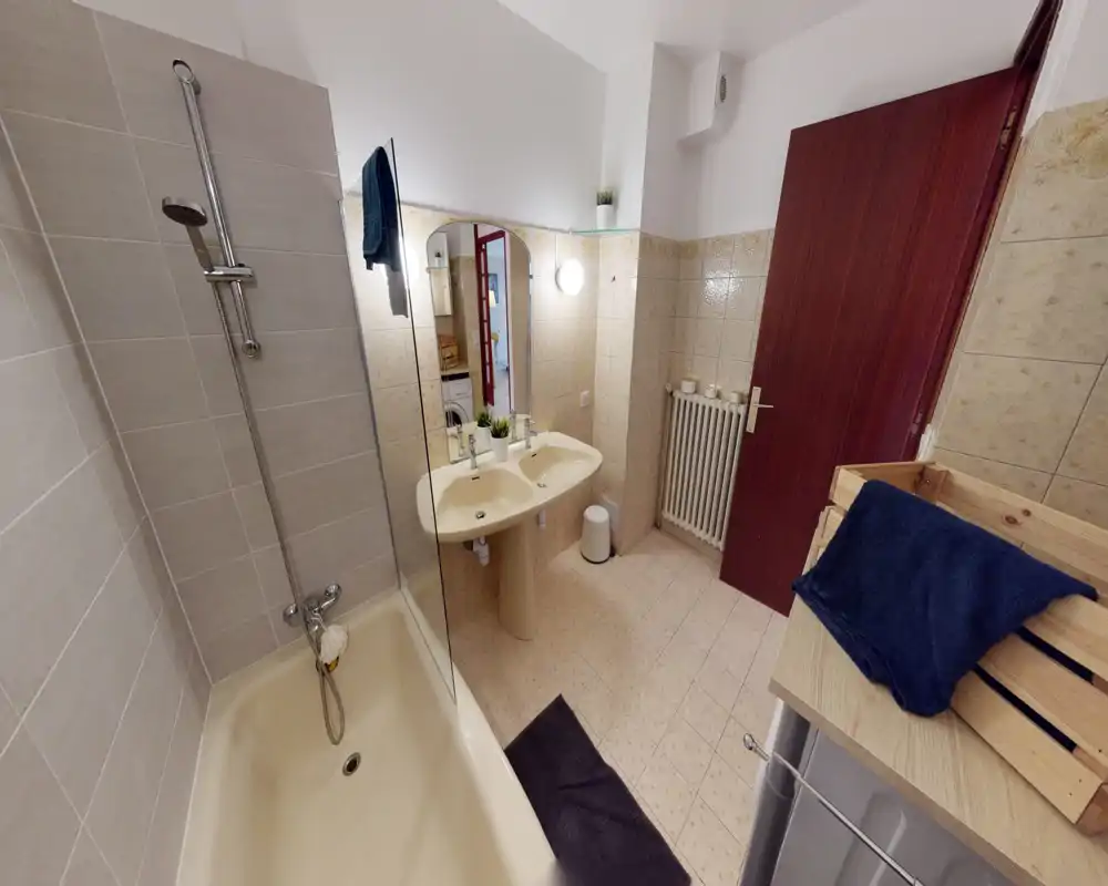 Clean, well-lit bathroom with a bathtub and wall-mounted shower, double sink, mirror and tiled walls — practical and functional for guests.