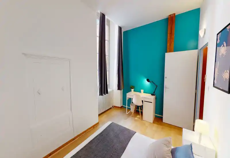 Chambre lumineuse et élégante avec un lit double au premier plan, un mur accent turquoise et un petit bureau près de la fenêtre. Grande fenêtre avec rideaux pour la lumière naturelle; le plancher en bois et les textiles rendent l'espace chaleureux.
