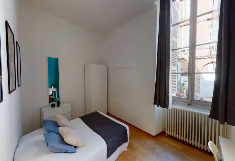 Chambre lumineuse au style épuré avec un lit double en linge neutre, coussins décoratifs et un plaid foncé ; grande fenêtre avec volets laissant entrer beaucoup de lumière et une armoire blanche pour le rangement.