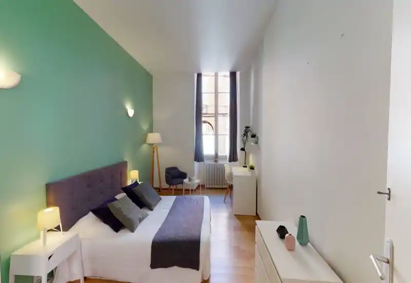 Chambre spacieuse et lumineuse avec un lit double confortable, tables de chevet et lampes, un fauteuil près de la fenêtre et un petit bureau — mur d'accent vert menthe et parquet chaleureux.