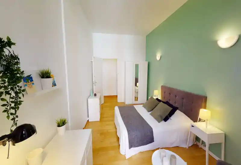 Chambre lumineuse et soignée avec un lit double confortable devant un mur vert, tables de chevet avec lampes et plantes décoratives pour une ambiance chaleureuse.