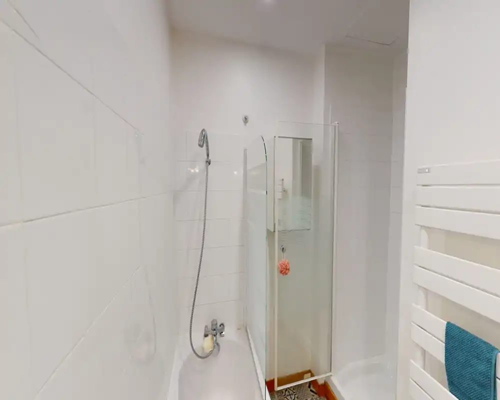 Salle de bains propre et compacte avec baignoire et douchette, cabine de douche avec écran en verre, murs carrelés blancs et sèche-serviettes — lumineuse et soignée.